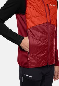 Gilet isolato rosso e arancione con motivi trapuntati, maglia a maniche lunghe nera, e dettaglio della tasca laterale con zip. Logo del marchio visibile.