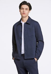 Giacca bottonata blu navy con colletto aperto, tasca frontale e texture liscia, abbinata a una maglietta bianca sotto e pantaloni coordinati.