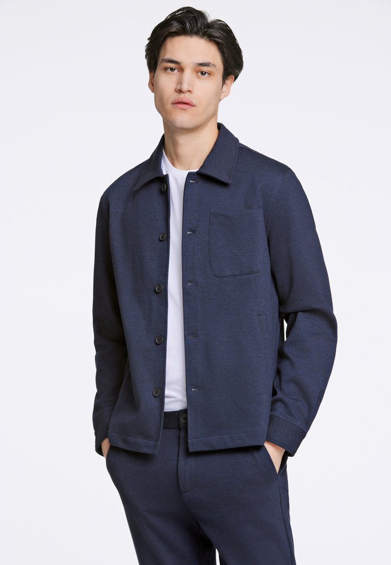 Giacca bottonata blu navy con colletto aperto, tasca frontale e texture liscia, abbinata a una maglietta bianca sotto e pantaloni coordinati.