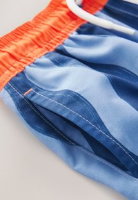 Costume da bagno blu con elastico arancione in vita, caratterizzato da motivi a onde, dettagli cuciti e coulisse. Realizzato in tessuto leggero.