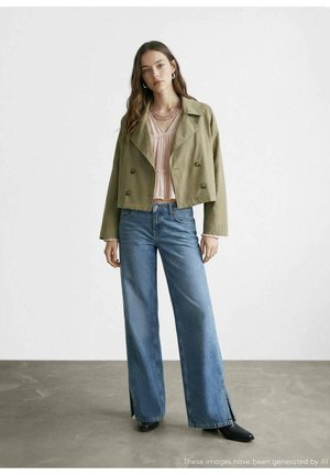 Mujer con chaqueta corta cruzada de color oliva, blusa rosa claro, vaqueros anchos con aberturas laterales y botines negros, de pie en el interior.