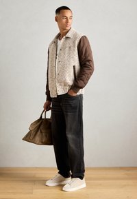 Giacca bomber grintosa con un corpo beige strutturato e maniche marroni imbottite, abbinata a jeans larghi e sneaker di colore chiaro. Porta una borsa marrone.