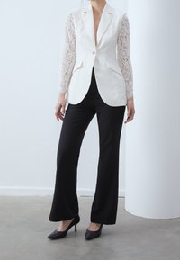 Biały żakiet z długimi koronkowymi rękawami, z pojedynczym zapięciem na guzik i dwoma przednimi kieszeniami, w połączeniu z czarnymi spodniami dzwonami i czarnymi szpilkami.