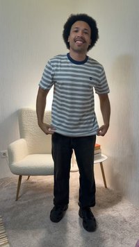 Mann med krøllete hår smiler, iført en hvit og marineblå stripet t-skjorte, svarte bukser og svarte sko, står i et rom med teppe og en lenestol.