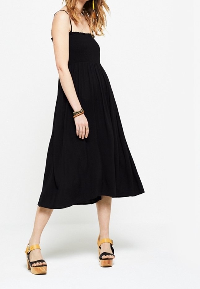 Femme portant une robe midi noire sans manches et des sandales compensées jaunes, debout devant un fond blanc.