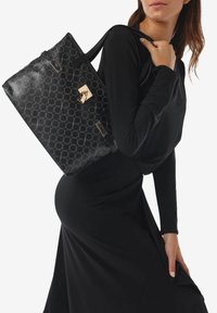 Borsa tote in pelle nera con un motivo geometrico, dettagli in metallo dorato e due manici robusti; mostrata su un modello che indossa un abito nero.