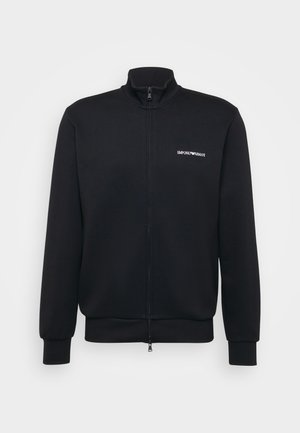Schwarze Zip-Up-Trainingsjacke aus weichem Stoff. Verfügt über einen hohen Kragen und gerippte Bündchen, mit einem weißen Logo auf der linken Brust.