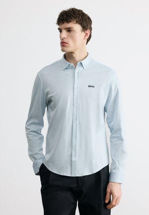 Młody mężczyzna w jasnoniebieskiej koszuli z guzikiem i logo "BOSS" na piersi, stojący z rękami w kieszeniach, patrzący w bok.