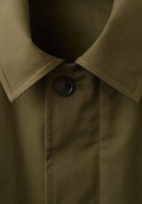 Primo piano del colletto di una camicia verde oliva con un unico bottone scuro allacciato in alto.