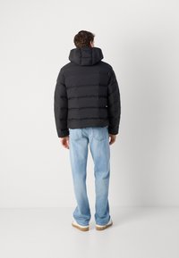 Zwarte pufferjack met een hoge kraag en capuchon, met horizontaal gequiltte secties. Draagt lichtblauwe jeans en tan schoenen.