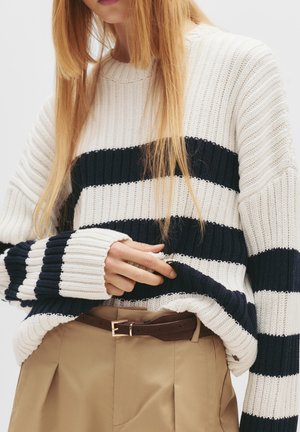 Sweter w paski w kolorze kremowym i granatowym, mający prążkowaną teksturę oraz szerokie rękawy, zestawiony z beżowymi spodniami i brązowym paskiem.