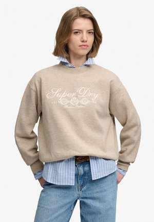Młoda osoba z krótkimi włosami nosi beżową bluzę SuperDry na niebieskiej koszuli w paski i niebieskich dżinsach, stoi z rękami w kieszeniach.