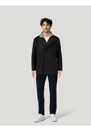 Un manteau noir avec un col à doublure grise, fermeture zippée unique et accents de boutons. Porté sur une chemise blanche et un pantalon marine. Chaussures blanches.