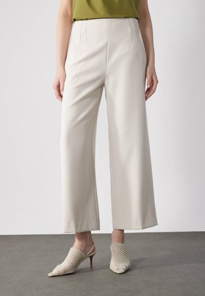 Vrouw draagt crèmekleurige, wijde cropped broek, groene top en crèmekleurige gevlochten pumps met hoge hak en slingback, staand op een grijze vloer.
