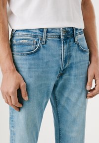 Világoskék farmernadrág, slim fit szabásban, öt zsebbel, fém szegecsekkel és egy címkével az első zseben. A textúra sima és enyhén kopott.