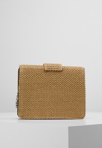 Obdélníková hnědá tkaná psaníčko s horní klopou a stříbrným řetízkem, vystavené na bílém podstavci na šedém pozadí.