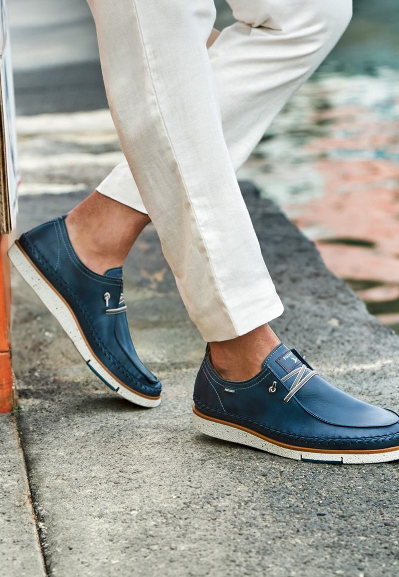 Homme portant des chaussures décontractées en cuir bleu avec des semelles blanches et un pantalon beige, debout sur une surface en béton près de l'eau.