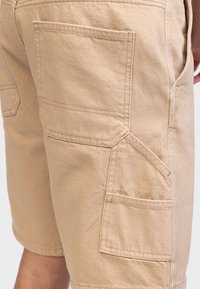 Beige Shorts mit mehreren aufgenähten Taschen an der Seite und am Rücken, getragen von einer Person mit leicht gebeugtem Arm, der sichtbar ist.
