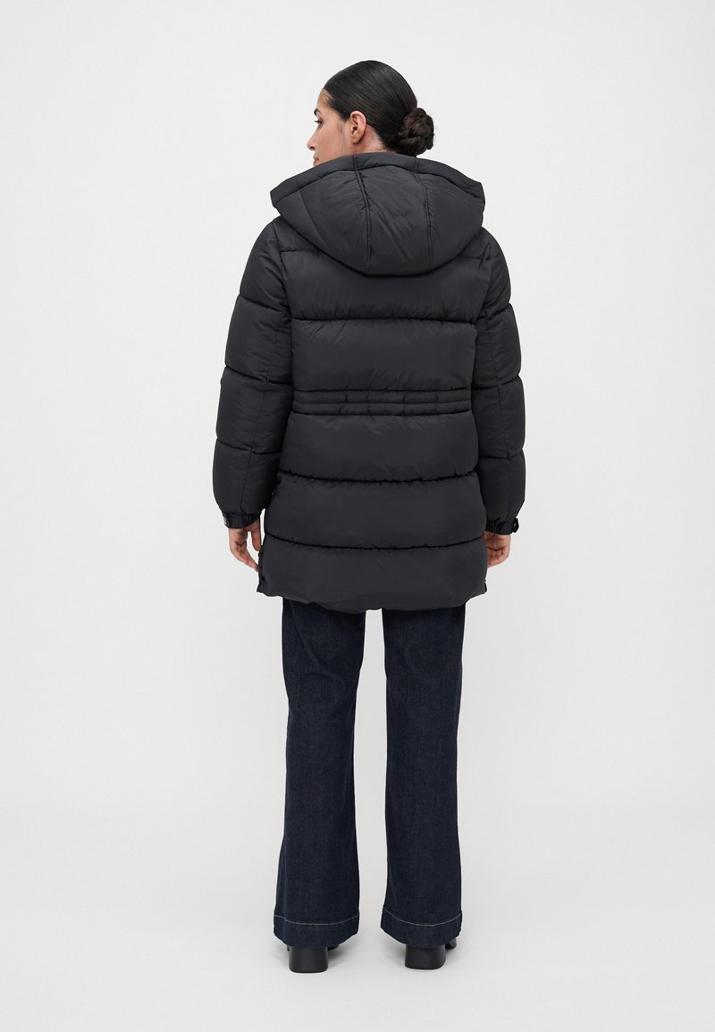 Mujer con el cabello oscuro recogido en un moño, vistiendo un abrigo de plumas negro con capucha y jeans oscuros de pierna ancha, vista desde la parte de atrás sobre un fondo blanco liso.