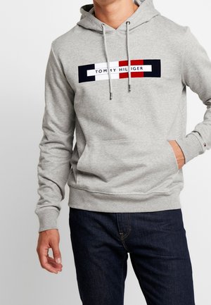 Sudadera gris con un parche de logo en el frente que presenta elementos en rojo, azul marino y blanco. Incluye capucha con cordón y bolsillo tipo canguro. Textura suave.