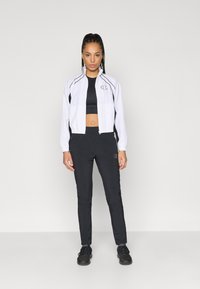 Balta užtrauktuku susegama striukė su juodais akcentais, derinama su juodu trumpu topu ir kelnėmis. Medžiaga atrodo lengva ir sportiška. Ant striukės ir kelnių yra logotipas.