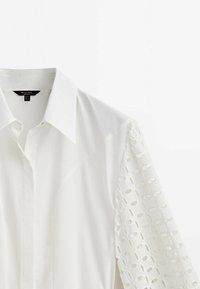 Blouse blanche avec col classique, présentant des détails en dentelle texturée sur les manches et un tissu lisse pour le corps. Manches courtes, design moderne.