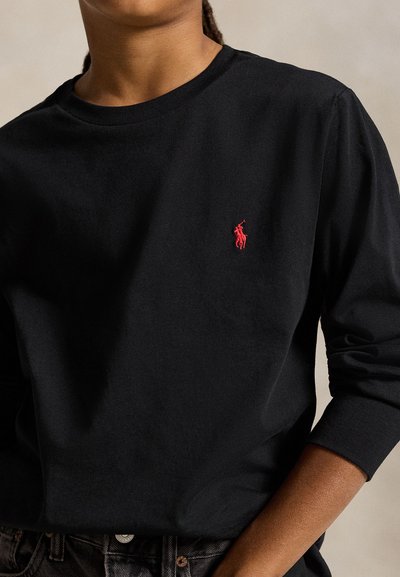 Person wearing a black long-sleeve shirt with a small red embroidered logo of a polo player on the left chest.