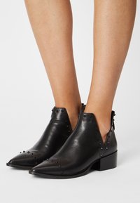 Stivaletti in pelle nera con punta appuntita, piccoli chiodini decorativi, tacco basso a blocco, indossati a piedi nudi su uno sfondo bianco.