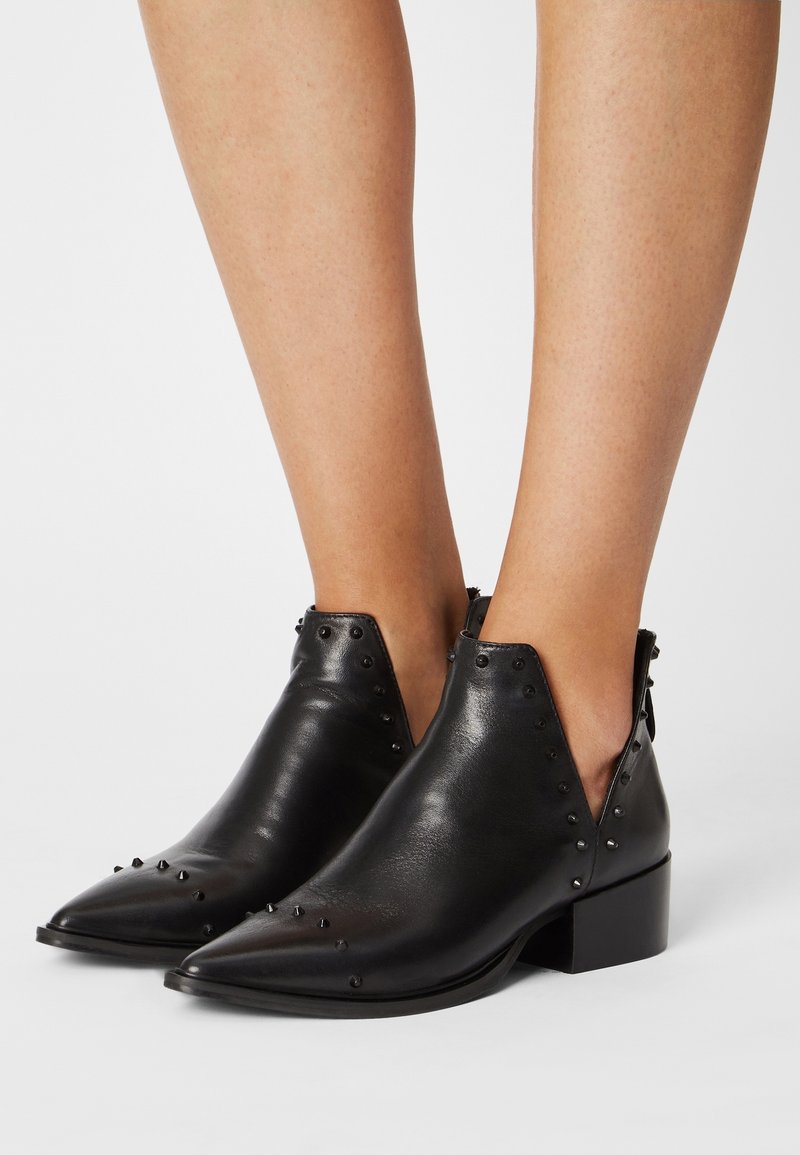 Stivaletti in pelle nera con punta appuntita, piccoli chiodini decorativi, tacco basso a blocco, indossati a piedi nudi su uno sfondo bianco.