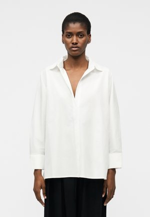 Kobieta stojąca na jednolitym tle, ubrana w luźną białą koszulę z guzikami, z rozpiętym kołnierzykiem, oraz czarne spodnie.