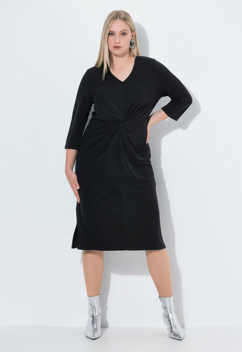 Vestido negro hasta la rodilla con mangas de tres cuartos, que presenta un detalle frontal retorcido y aberturas laterales. La modelo lleva botas de tobillo plateadas.