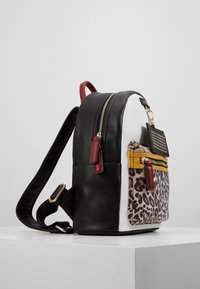 Mochila pequeña con paneles en negro, blanco y estampado de leopardo, bolsillo con cremallera color mostaza, tiradores de cremallera rojos y una etiqueta negra con tachuelas adjunta.