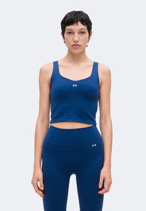 Femme aux cheveux courts et foncés portant un crop top bleu sportif et des leggings taille haute assortis avec les logos blancs Under Armour.