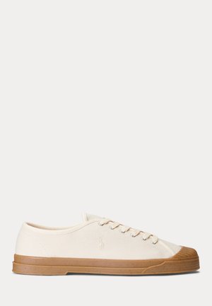 Lavtop beige lærredssneaker med brun gummisål og tåhætte, snørelukning, side logo, vist mod en ensfarvet lys baggrund.