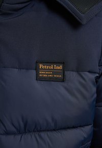 Jachetă puffer bleumarin cu o textură de țesătură netedă, cusături orizontale și o etichetă logo portocalie cu brandingul "Petrol Ind".