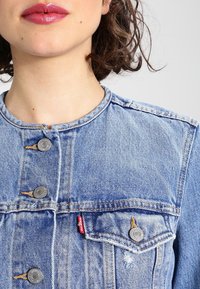 Světle modrá džínsová bunda s kulatým výstřihem, stříbrnými knoflíky, viditelným šitím a přední kapsou s červeným štítkem jako akcentem.