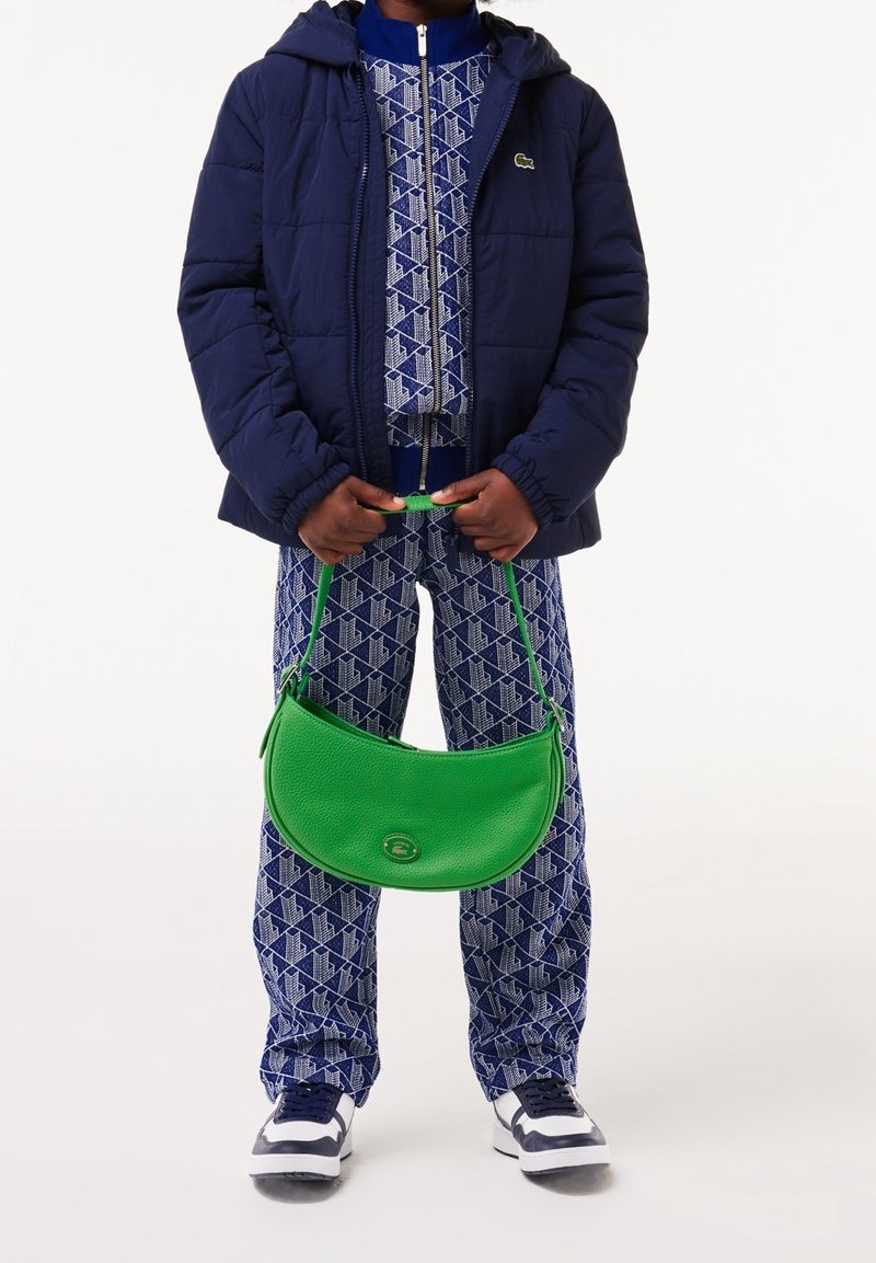 Green leather handbag with a curved shape and logo detail, worn with a navy quilted jacket and patterned blue pants.