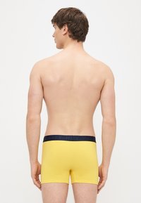 Sárga pamut férfi boxer, sötétkék derékpánttal, amelyen "HAPPY" felirat található. A fazon testhez simuló, a felső combot fedi, és sima tapintású.