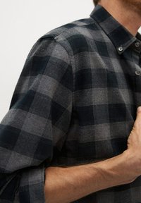 Chemise en flanelle présentant un motif à carreaux gris foncé et noir, un col à boutons et des manches retroussées, avec une texture douce.