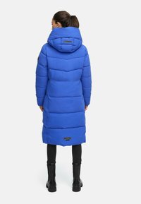 Lang blauwe pufferjas met een capuchon, voorzien van horizontale naden, textielstof en een logopatch op de achterkant. Gemaakt in combinatie met zwarte leggings en laarzen.