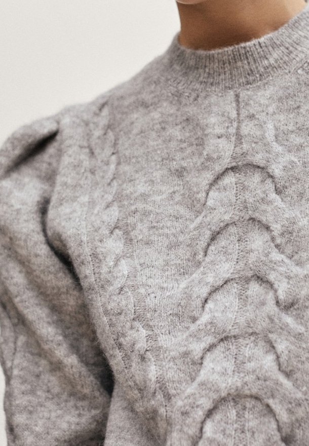Gros plan d'une personne portant un pull en maille gris clair avec des motifs torsadés et des épaules bouffantes.