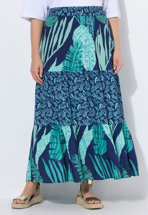 Frau trägt einen langen, gestuften marineblauen Rock mit grünen und türkisfarbenen tropischen Blattmustern, kombiniert mit goldenen Keilsandalen.