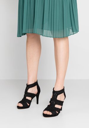 Sandalias de tacón alto negras con tiras trenzadas, punta abierta y tacón puntiagudo. El material parece suave con detalles texturizados y acentos.