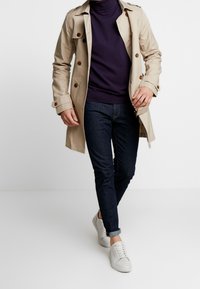 Beżowy płaszcz trench z brązowymi guzikami na fioletowym golfie, zestawiony z ciemnymi dżinsami i białymi sneakerami. Modelka przedstawiona w pozie podczas chodzenia.