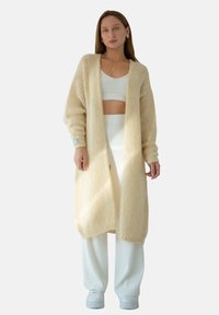 Długi, jasnobeżowy sweter o dzianinowej strukturze z prążkowanym teksturem i opuszczonymi ramionami, zestawiony z białym crop topem i szerokimi spodniami.