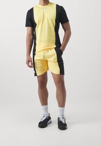 Camiseta atlética de manga corta amarilla y negra con pantalones cortos negros. Presenta el logo de Adidas. Se lleva con zapatillas negras y calcetines blancos. Diseño limpio.