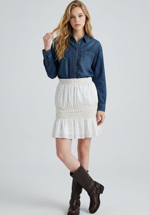 Mujer rubia con una camisa vaquera oscura, falda blanca con detalles de encaje y botas altas marrón oscuro, de pie con una pierna cruzada delante.
