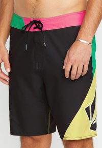 Shorts de bain avec un corps noir, une taille vert et rose, des accents jaunes et un motif de design distinctif sur le côté.