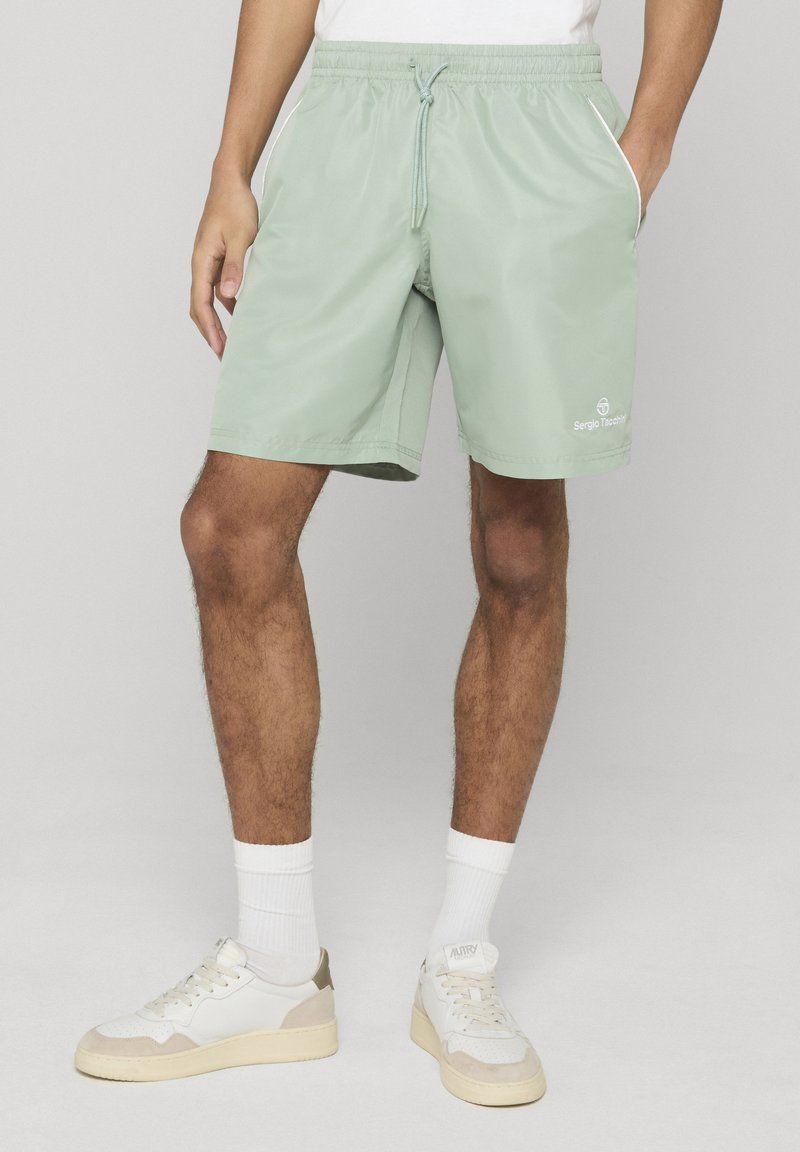 Homme portant un short à cordon vert menthe, des baskets blanches, des chaussettes et une chemise blanche, debout avec une main dans la poche.