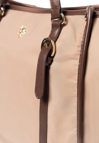Borsa in tessuto beige con dettagli in pelle marrone, accessori in oro e forma strutturata. Presenta una texture liscia e un logo decorativo.